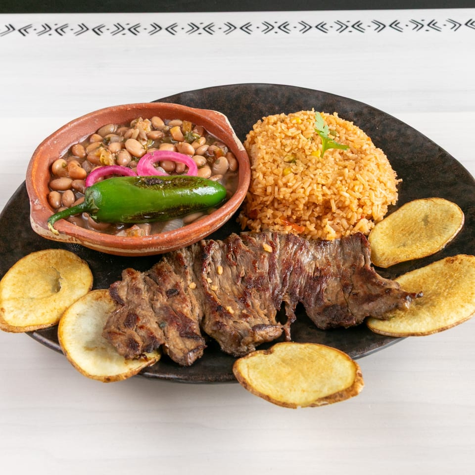 Grilled steak, rice, beans with a green pepper, and potato slices on a dark plate.