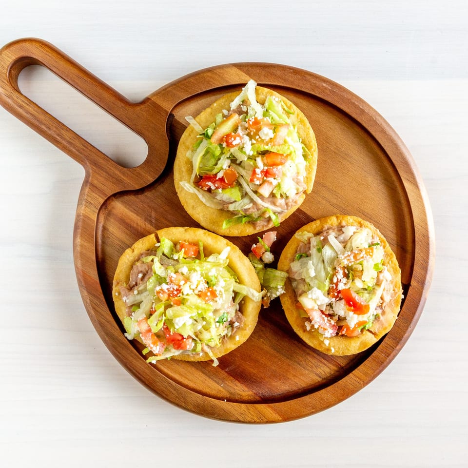 Three sopes with refried beans, shredded lettuce, diced tomatoes, and crumbled cheese on a wooden board.