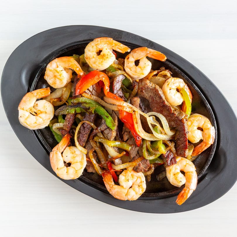 Sizzling shrimp and steak fajitas with colorful bell peppers and onions on a black platter.
