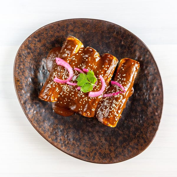 Three enchiladas covered in dark mole sauce, topped with sesame seeds, pickled red onions, and cilantro.