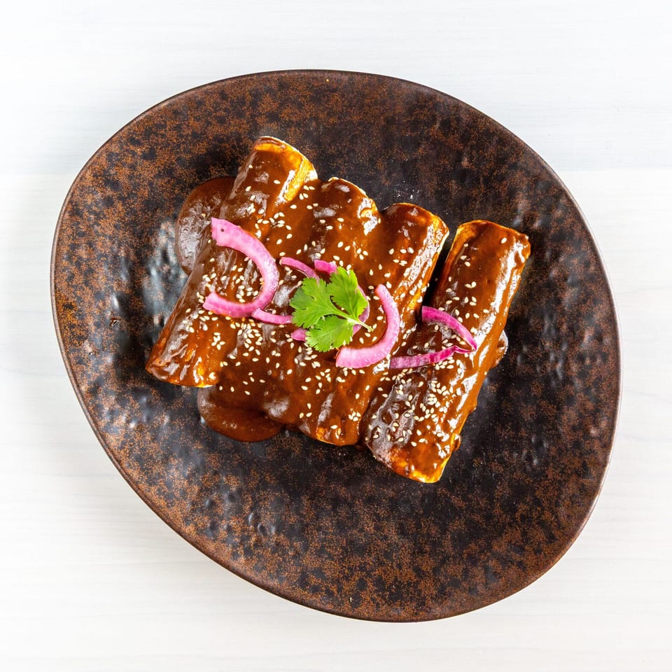 Three enchiladas covered in dark mole sauce, topped with sesame seeds, pickled red onions, and cilantro.