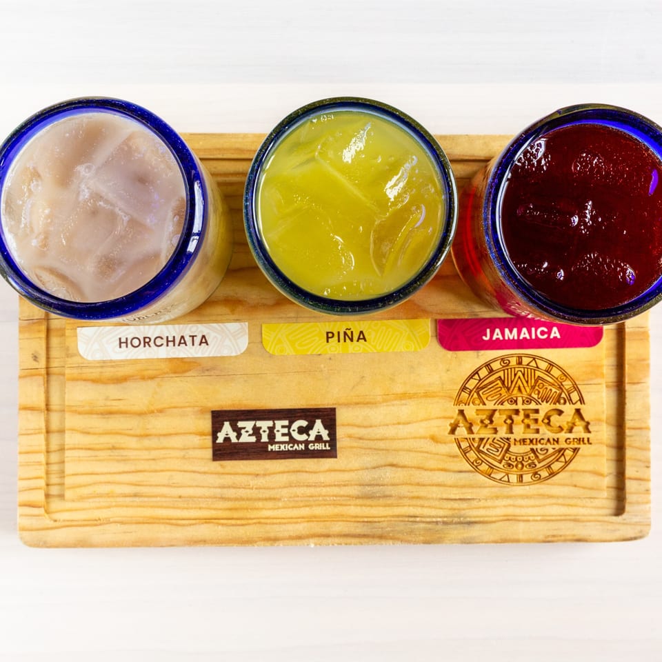 Three colorful Mexican drinks, Horchata, Piña, and Jamaica, on a wooden serving board from Azteca Mexican Grill.