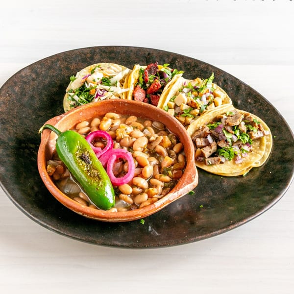 A black plate holds a bowl of pinto beans with a grilled jalapeño, surrounded by four diverse tacos.