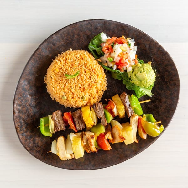 Two skewers of grilled meat and vegetables, with rice, pico de gallo, and guacamole on a dark plate.