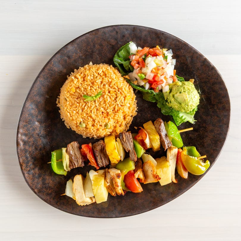 Two skewers of grilled meat and vegetables, with rice, pico de gallo, and guacamole on a dark plate.